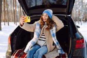 Young woman sitting in the trunk of her car taking a selfie against the backdrop of a snowy winter forest.